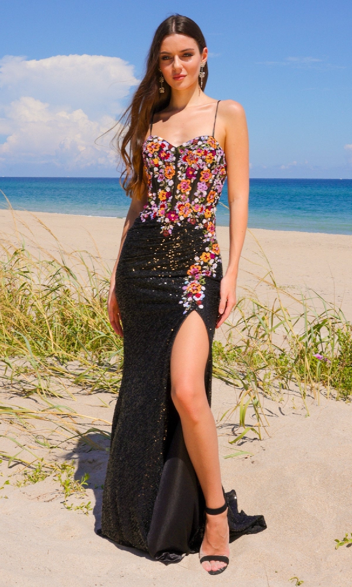 A woman stands on a sandy beach in the Multicolored Sequin Prom Gown: Amelia Couture 7061, featuring a stunning floral bodice and thigh-high slit, with the ocean and blue sky as a dreamy backdrop.